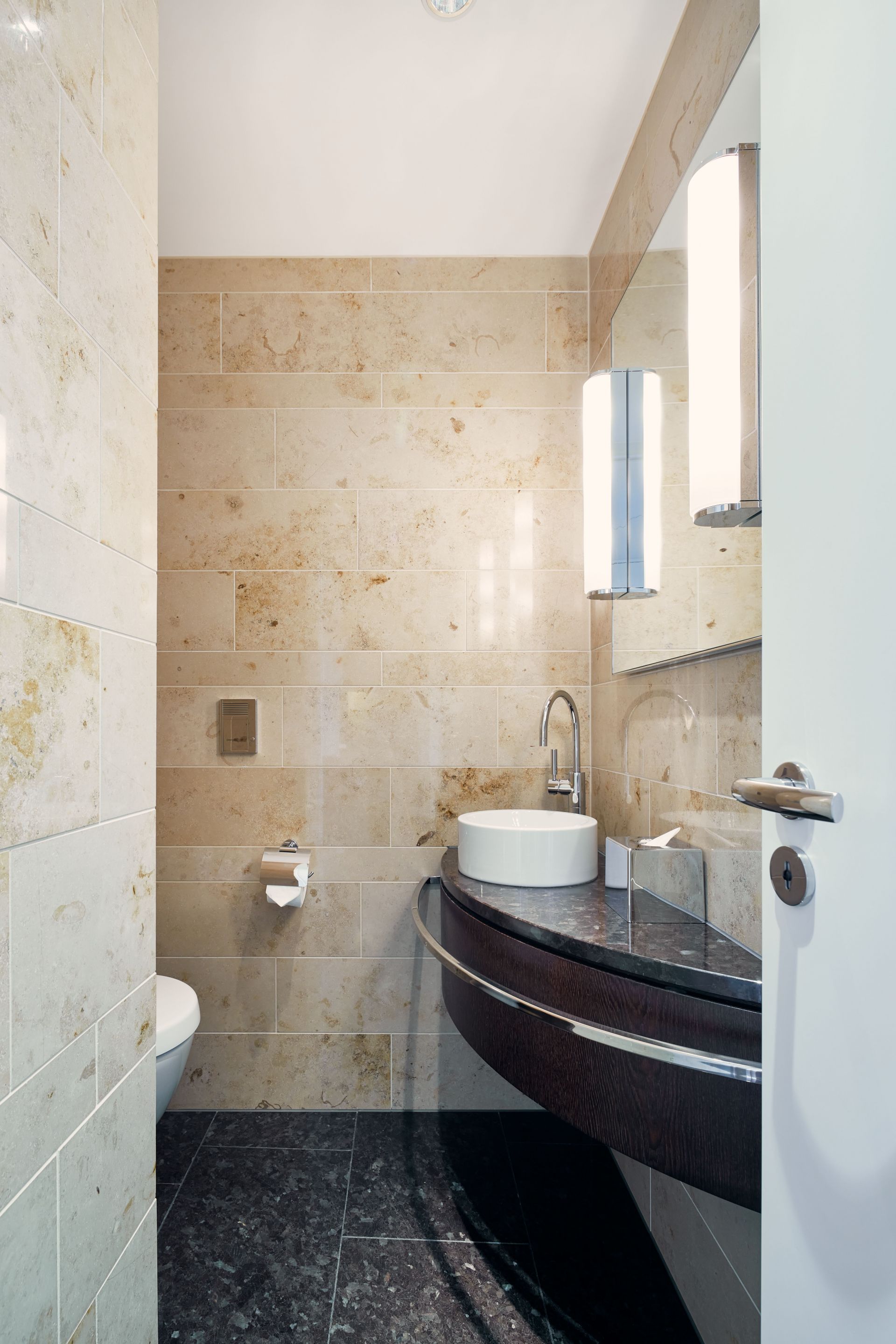 Ein modernes, kompaktes Badezimmer in der Golf Suite Dolder Grand verfügt über beige Marmorwände, einen dunklen Marmorboden, eine wandmontierte Toilette, ein rundes Aufsatzwaschbecken auf einer geschwungenen Ablage, einen Wandspiegel und elegante vertikale Leuchten.