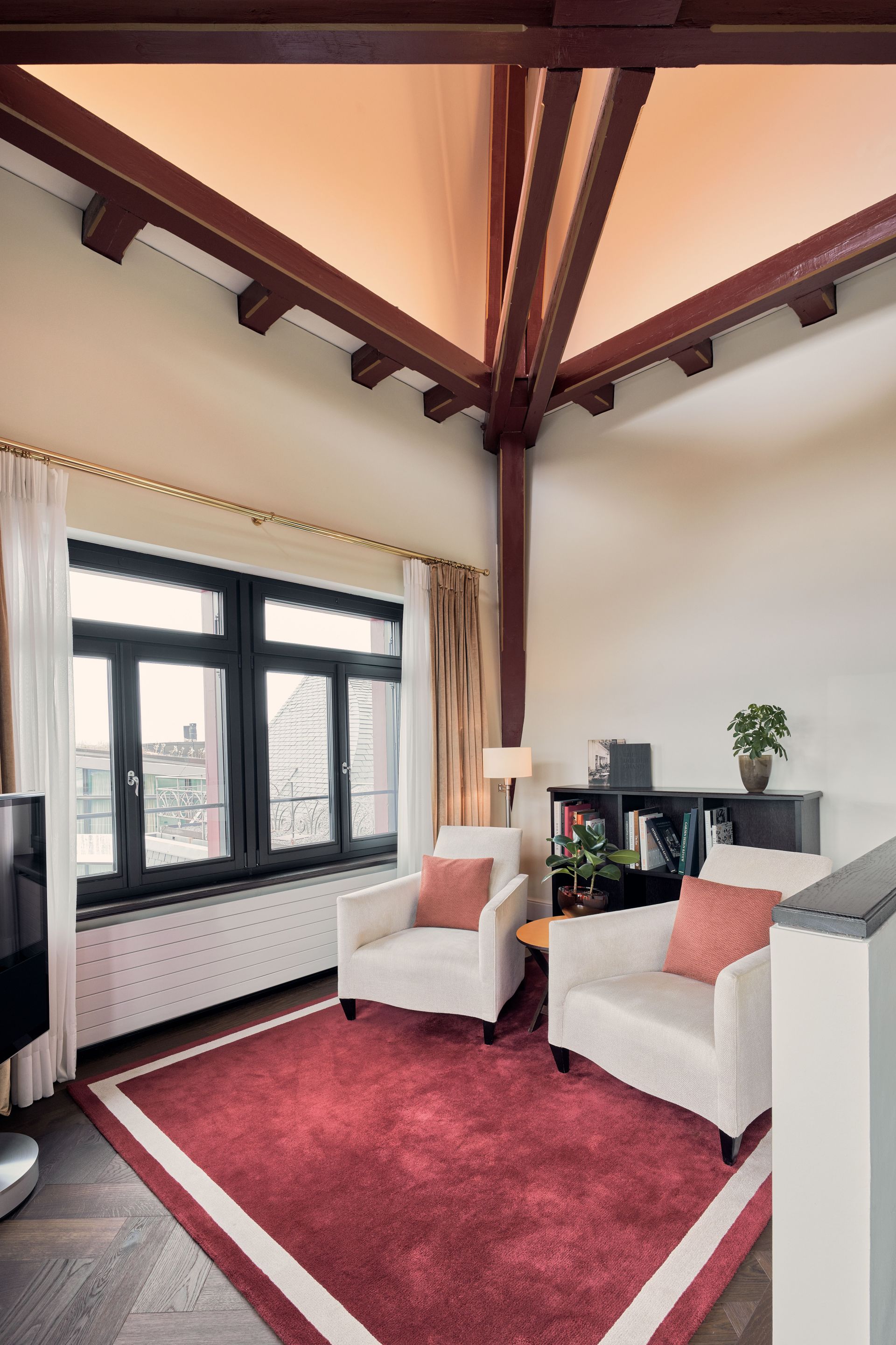 Ein gemütliches Wohnzimmer in der Suite Grand im Dolder Grand mit zwei cremefarbenen Sesseln, roten Akzentkissen, einem roten Teppich, großen Fenstern mit beigen Vorhängen, einem schwarzen Bücherregal, einem kleinen Tisch mit einer Lampe und freiliegenden roten Deckenbalken.